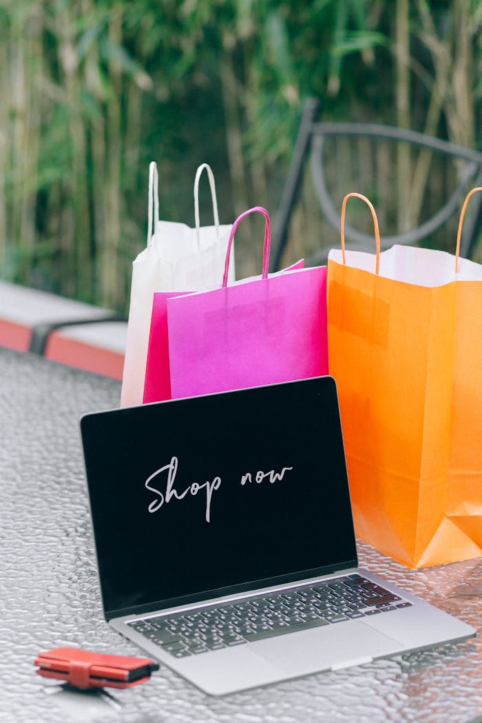 Laptop displaying 'Shop now' with vibrant shopping bags on glass table outdoors.